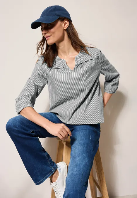 Frau mit blauer Kappe, gestreiftem Oberteil und Jeans sitzt auf einem Hocker.